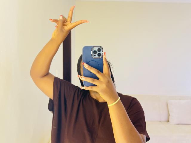 ThickMari met een bruin shirt, die een blauwe telefoon vasthoudt, met haar rechterarm omhoog, met oranje nagels, een gele polsband, een zichtbare iPhone-camera, een spiegelzelfportret, donker haar, bruine huid..