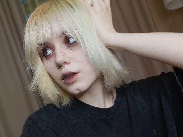 YasukoCella has blonde bob, freckles, nose ring, and dark makeup. She's looking up with hand in hair, wearing a black shirt, and has a slight pout.