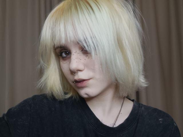 YasukoCella with platinum blonde, straight bob, wearing a black shirt, nose ring, lip piercing, freckles, and a silver necklace, looking slightly to the side with a neutral expression.