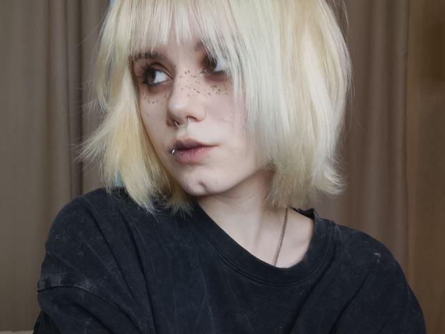 YasukoCella tiene el cabello rubio, corto y con flequillo, una camisa negra, un piercing en la nariz y un piercing en el labio. Tiene pecas en su rostro pálido y mira hacia un lado con una expresión seria..