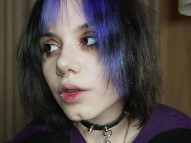 YasukoCella tiene la piel pálida, el cabello negro y morado, un piercing en la nariz, un piercing en el labio y un collar negro con un adorno. Mira hacia un lado con ojos azules..