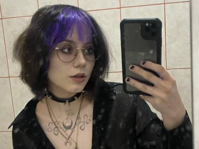 YasukoCella mit lila und schwarzen Haaren, Brille, schwarzem Choker, schwarzem Hemd, Halskette und Tattoo, macht ein Selfie vor einem Spiegel und zeigt ihre Brüste, trägt schwarzen Nagellack, im Badezimmer..
