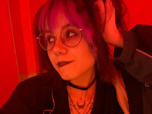 YasukoCella with purple and red hair, round glasses, black choker, dark lipstick, nose ring, and tattoos, holding hair, red-lit face, black jacket, and a confident expression.