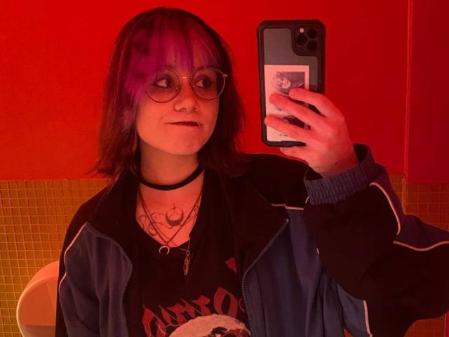 YasukoCella mit lila gefärbtem Haar, Brille, schwarzem Choker, mehreren Ketten, einem schwarzen Hemd und einer blauen Jacke, die ein Spiegel-Selfie in einem Badezimmer mit roten und gelben Fliesen macht..