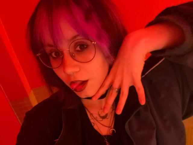 YasukoCella with purple hair, glasses, and red lipstick, sticking out tongue, making a rock gesture with finger, wearing black jacket, red light.