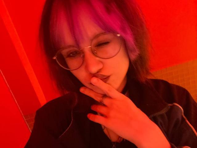 Yasuko, con capelli viola e neri, occhiali e un piercing al naso, che si morde un dito con un dito sulle labbra, indossa una giacca nera, illuminazione rossa..