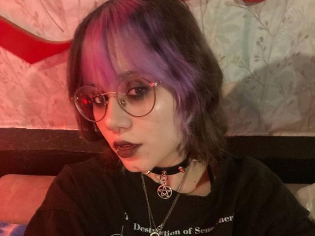 YasukoCella with purple and black hair, glasses, dark lipstick, black choker, pentagram necklace, black shirt, red and pink lighting, short wavy hair, looking at camera.