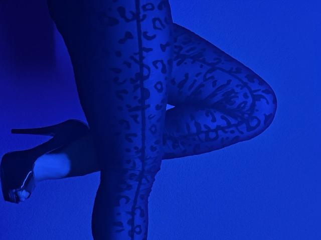 Le gambe di BalletSwan, indossanti collant a motivi blu, sono in posizione eretta su una gamba sola, con un tacco alto nero e il piede sollevato, illuminate da una luce blu..
