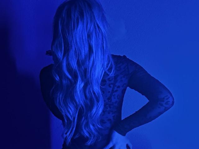 BalletSwan, avec de longs cheveux blonds ondulés, dos à la caméra, portant un haut en dentelle noire, les mains sur les hanches, sous un éclairage bleu. Ses fesses sont visibles, légèrement mises en valeur..