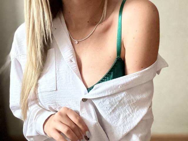 BalletSwan, avec ses cheveux blonds, porte une chemise blanche à une épaule qui révèle un soutien-gorge vert, un collier fin, et a une peau claire, exposant ses seins. Elle tient la chemise d'une main..