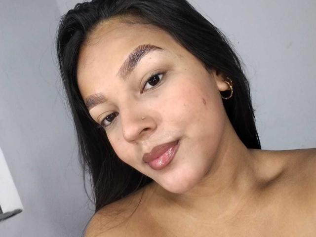 SheilaBrunette has tan skin, black hair, a nose piercing, and is smiling with glossy pink lips. She's wearing gold hoop earrings and has a small zit on her cheek.