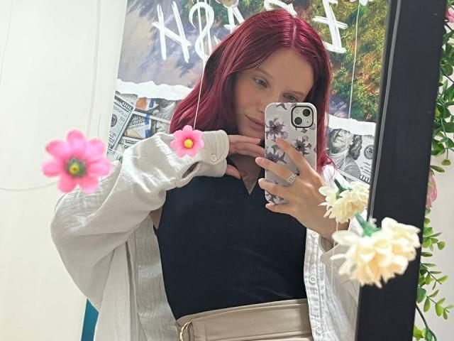 JuliannaMoor with red hair, black top, beige belt, white cardigan, holding phone, pink and white flowers in mirror, ring on finger, reflection, phone case with floral design.