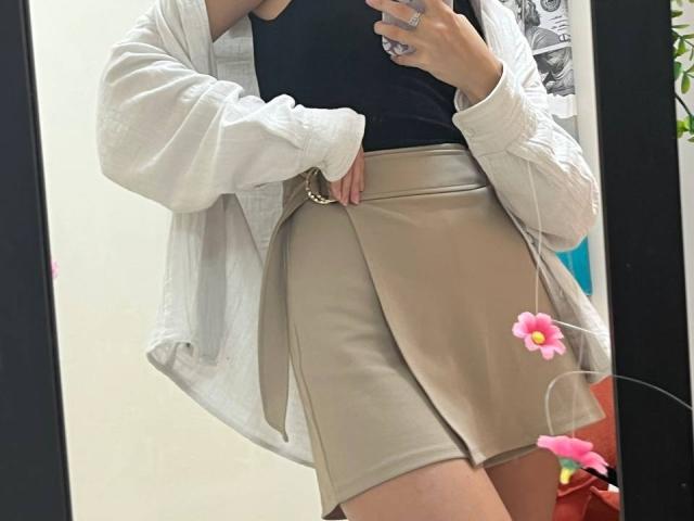 JuliannaMoor in a beige skirt, black top, white jacket, holding phone, mirror selfie, showing her ass, pink flower filter, ring on finger, reflection, partial face not visible.