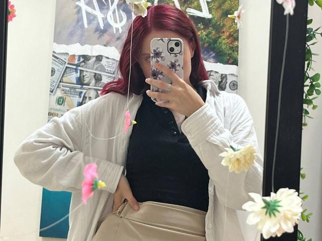 JuliannaMoor with red hair, wearing a black top and beige cardigan, taking a mirror selfie, showing her phone with floral cases, her hand on her hip, and flowers in mirror.