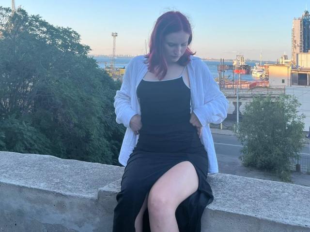 JuliannaMoor with red hair, wearing a black dress and white cardigan, sitting on a concrete ledge, pulling up her dress to reveal her thigh, looking down.