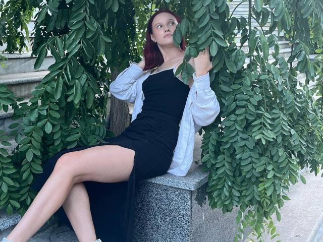 JuliannaMoor with red hair, black dress, white cardigan, sitting on a stone bench, legs crossed, hands in leaves, green foliage around, sunlight, serious expression, bare legs, pretty.