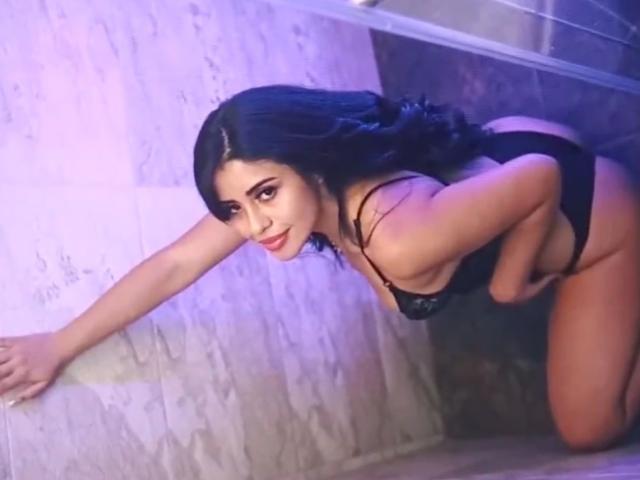 AgathaVictoria on her knees, leaning forward with hands on a wall, wearing black lingerie, showing her ass and small tits, with long dark hair and a seductive smile.