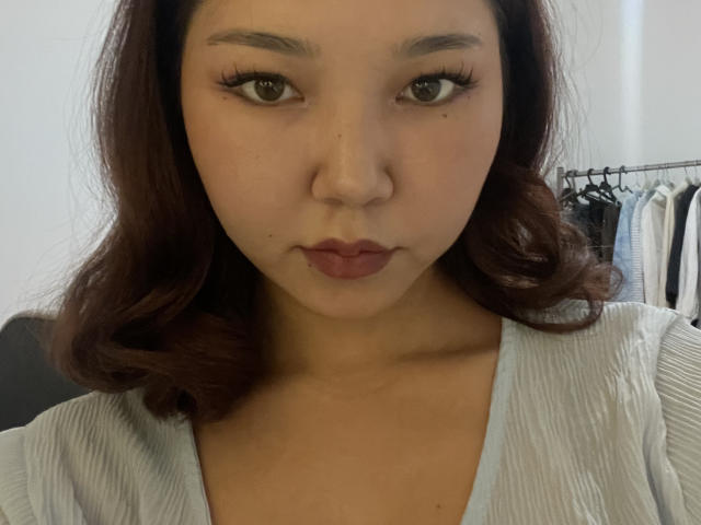 ElbaHuppenbauer heeft bruine ogen, volle roze-rode lippen en golvend donkerbruin haar. Ze draagt een lichtgrijze top met een V-hals en kijkt recht in de camera..