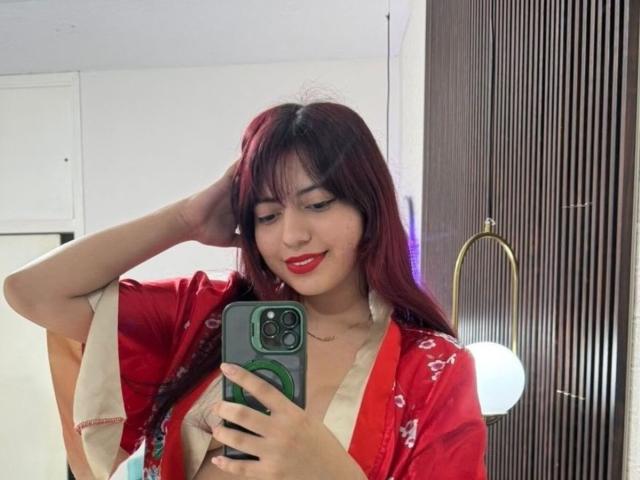 BrendaaPierce with red lipstick, red and white kimono, holding green phone, smiling, red hair, right hand in hair, white wall, brown vertical slats, standing, mirror selfie.