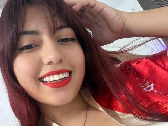 BrendaaPierce with red lipstick, smiling, showing white teeth, hand in hair, wearing red shiny top, has long red hair, light brown skin, green eyes, and a thin necklace.