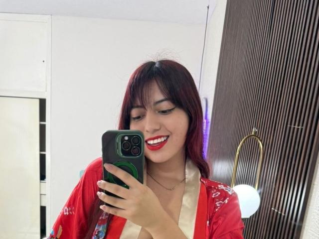 BrendaaPierce with red hair, red lipstick, and red floral top, holding a green and black phone, smiling, showing her tits slightly, in a mirror selfie.