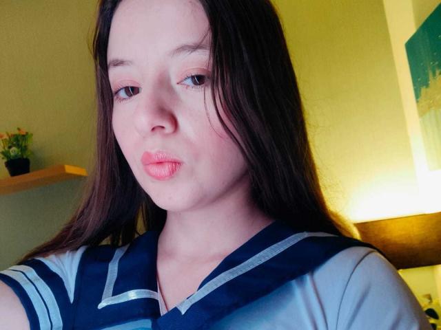 VioletaNova with puffy pink lips, long brown hair, and a blue and white sailor-style shirt, looking slightly bored. Her small, fair-skinned face is centered in the shot.