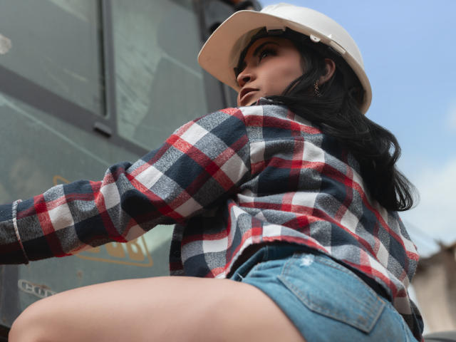 Alana West, con cabello oscuro, usando un sombrero blanco, una camisa de cuadros rojos y azules, y pantalones cortos vaqueros, mostrando su trasero mientras mira por encima del hombro..