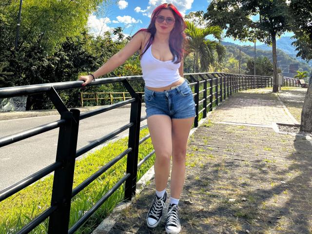 MaraCortes met rood haar, een witte tanktop en denim shorts, leunt tegen een zwarte reling, draagt zwarte en witte sneakers, en laat haar borsten zien, in een zonnige, open omgeving..