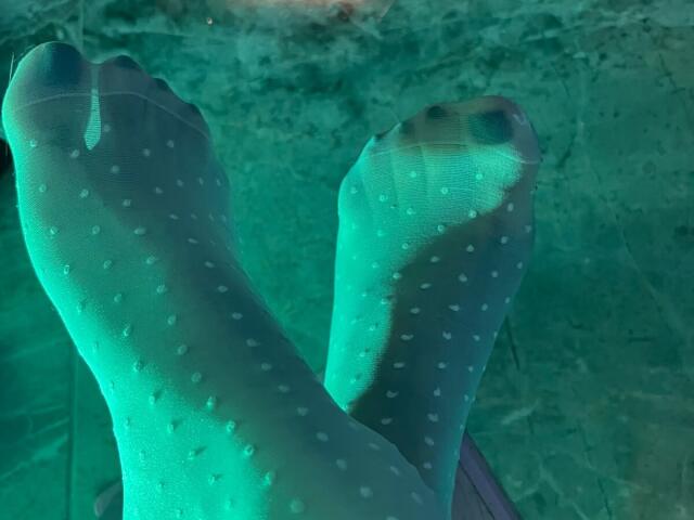 Les pieds de LiaMiss, portant des chaussettes vertes à pois semi-transparentes, laissant apparaître ses orteils, sont éclairés par une lumière verte. Ses orteils sont légèrement recourbés et les chaussettes sont légèrement froissées..