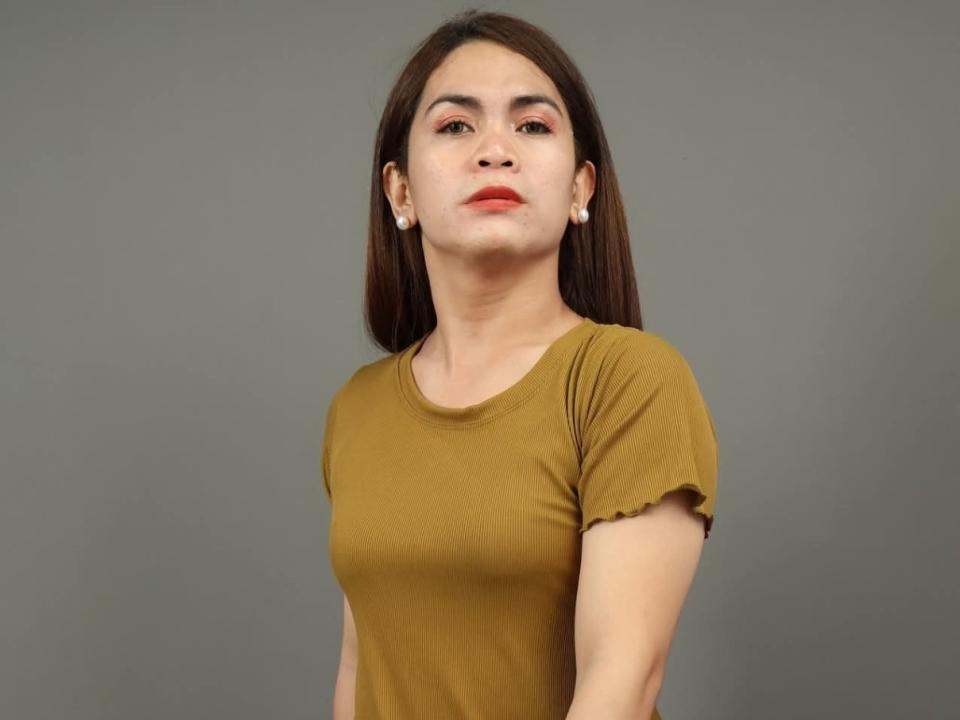 JanellaTs with brown hair, red lipstick, small white earrings, and a mustard t-shirt, looking slightly up with a neutral expression. Her tits are small and her ass is not visible.
