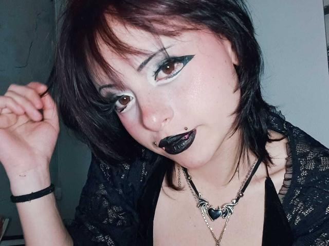 ViolettClay ha un rossetto nero scuro, un trucco occhi con ali nere ben definite, una camicia di pizzo nero, un anello al naso e una collana a forma di cuore, con i capelli castani corti e un'espressione giocosa..