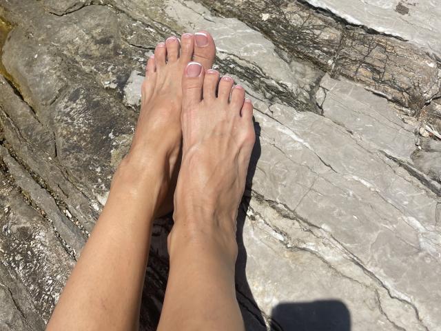 SandraSweet's bare feet with pink toenails resting on rough, gray stone. Her skin is tan, and the stone has dark, jagged lines. Sunlight casts a shadow.