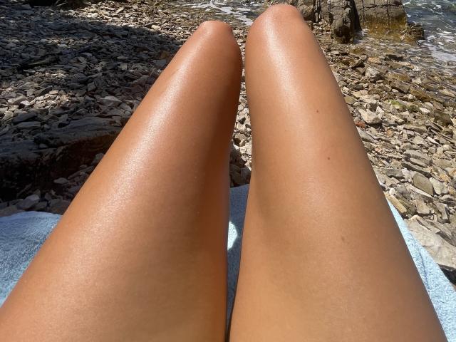 SandraSwweet's shiny, tan, bare-assed legs are in the foreground, pointing slightly apart, with a rocky, pebbly shoreline visible in the background under bright sunlight.