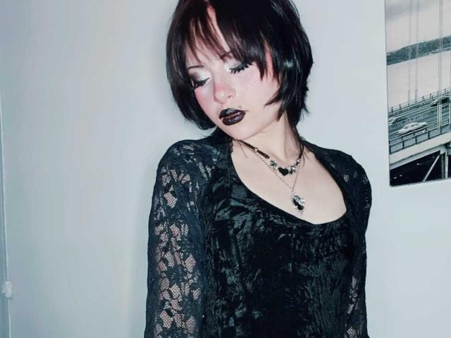 ViolettClay, con i capelli neri corti, indossa una camicetta di pizzo nero, rossetto scuro e due collane, in piedi contro un muro bianco, con lo sguardo rivolto verso il basso e un'espressione leggermente malinconica..