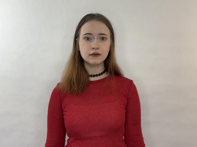Marita Pavelko, con cabello castaño lacio, vestida con una camisa roja de manga larga, un collar negro y lápiz labial rojo, está de pie frente a un fondo blanco liso. Los senos son pequeños, el rostro es neutro..