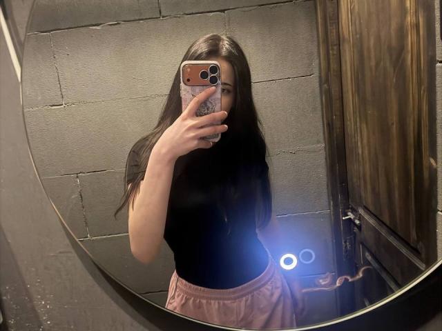 LienDepippo with long dark hair, black top, pink skirt, holding phone in mirror selfie, showing slim body, small tits, in dimly lit room, using ring light.