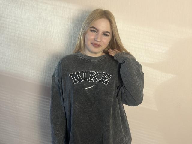 MariluChouinard with long blonde hair, wearing a gray Nike sweatshirt, standing against a wall, hand near her face, smiling slightly, subtle shadow on the right.