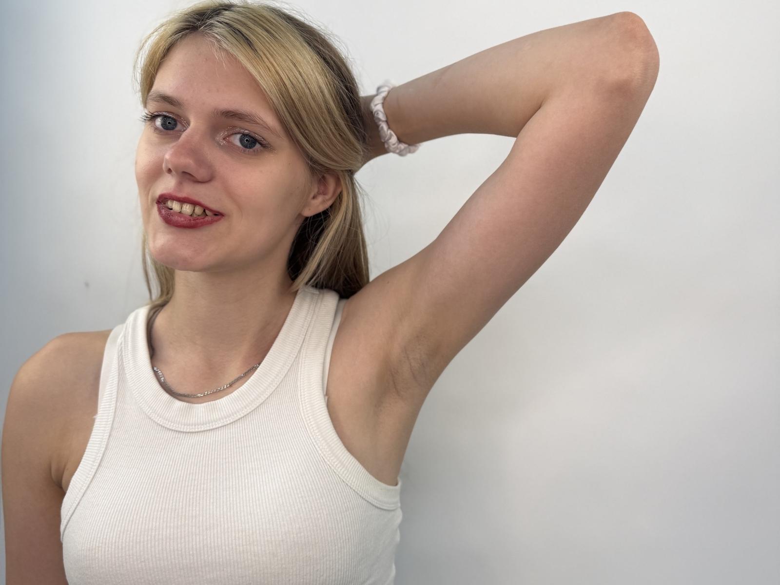 FloreneMcclendon has blonde hair, blue eyes, and red lipstick, wearing a white tank top, arm raised, showing armpit hair, with a white scrunchie on her wrist.