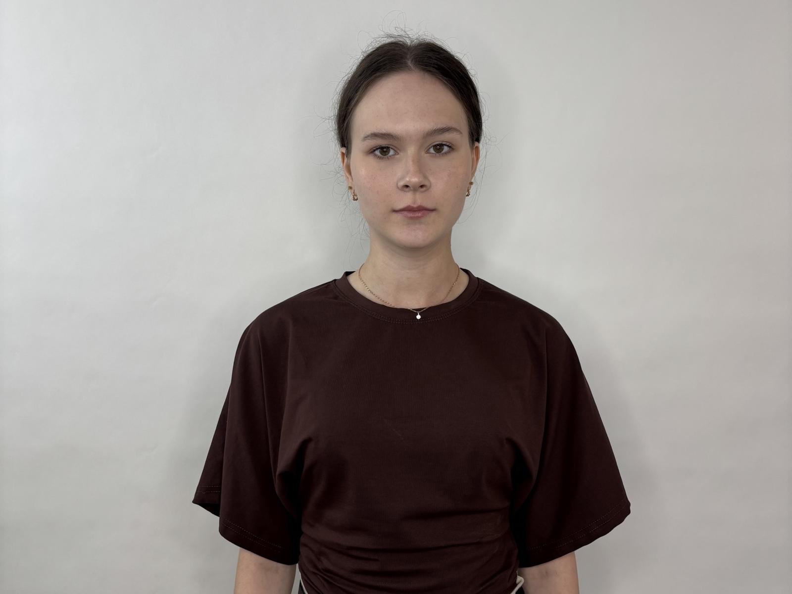 Anastacia Daquino mit glattem, dunklem Haar, die ein braunes Hemd trägt und vor einer schlichten weißen Wand steht, blickt direkt in die Kamera mit einem neutralen Gesichtsausdruck, kleine Ohrringe sind sichtbar..
