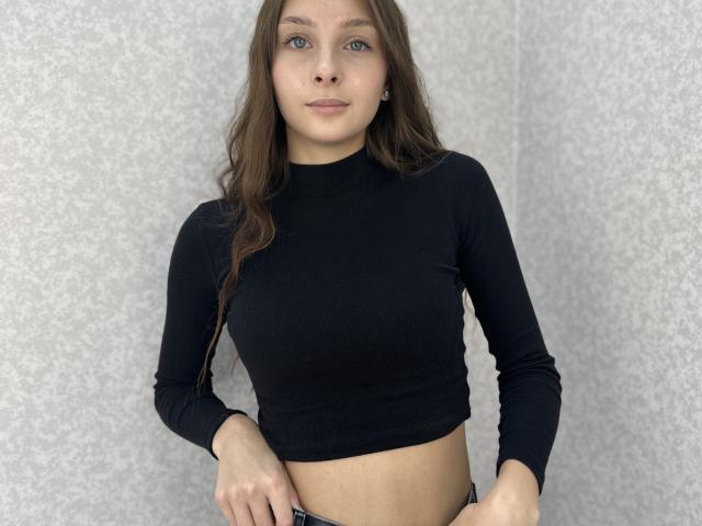 Dottie Larson, con lunghi capelli castani, occhi azzurri e piccoli seni, indossa una crop top nera a maniche lunghe, ed è in piedi con le mani sui jeans, di fronte a una parete grigia con una texture..