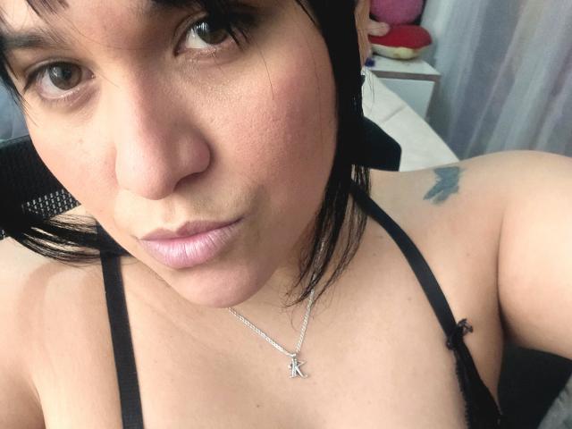 VivianaThompson has light brown skin, black hair, and a tattoo on her right shoulder. She's wearing a black strap top with a silver cross necklace, showing her tits.