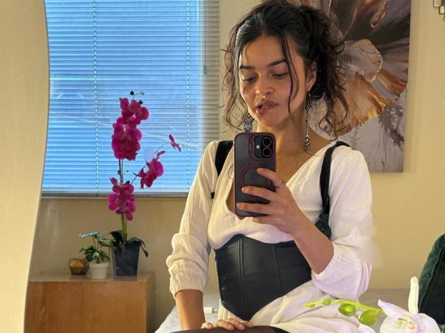 LucyySky with dark curly hair, white blouse, black corset, holding phone, taking mirror selfie, pink orchid, green leaves, window blinds, earrings, slightly pouty expression, subtle makeup.