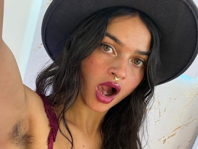 LucyySky with dark hair, brown eyes, and a nose ring, wearing a black hat and pink lipstick, open mouth, showing her pink tongue, and armpit hair visible.