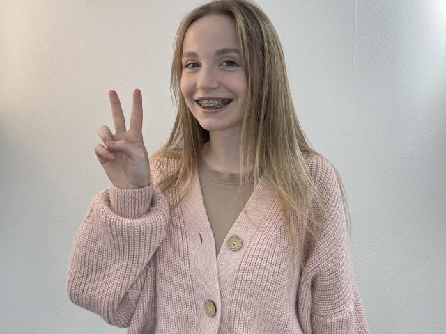 RubyAverill, blonde, smiling, wearing pink cardigan, beige shirt, holding up peace sign, metal teeth, cute, white wall background, casual, soft light, relaxed, gentle expression.
