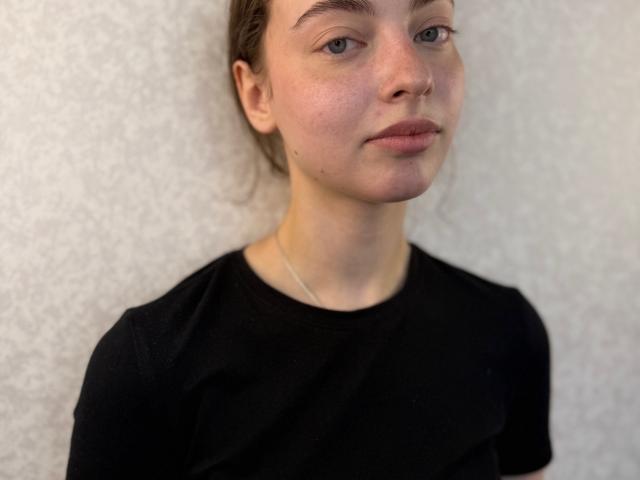 CharisseChiapetto has light skin, blue eyes, and brown hair in a messy bun. She wears a black shirt and a thin necklace, with a neutral, slightly smug expression.