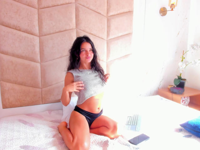 LucyySky kneels on a bed, lifting her gray crop top to reveal her small tits, wearing black panties. Her dark hair is wavy, and she's smiling. A laptop and phone are nearby.