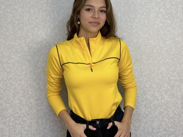 DremaSunday with wavy brown hair, wearing a yellow ribbed zip-up shirt and black pants, hands in pockets, smiling slightly, medium-sized tits, light skin, standing against a gray wall.