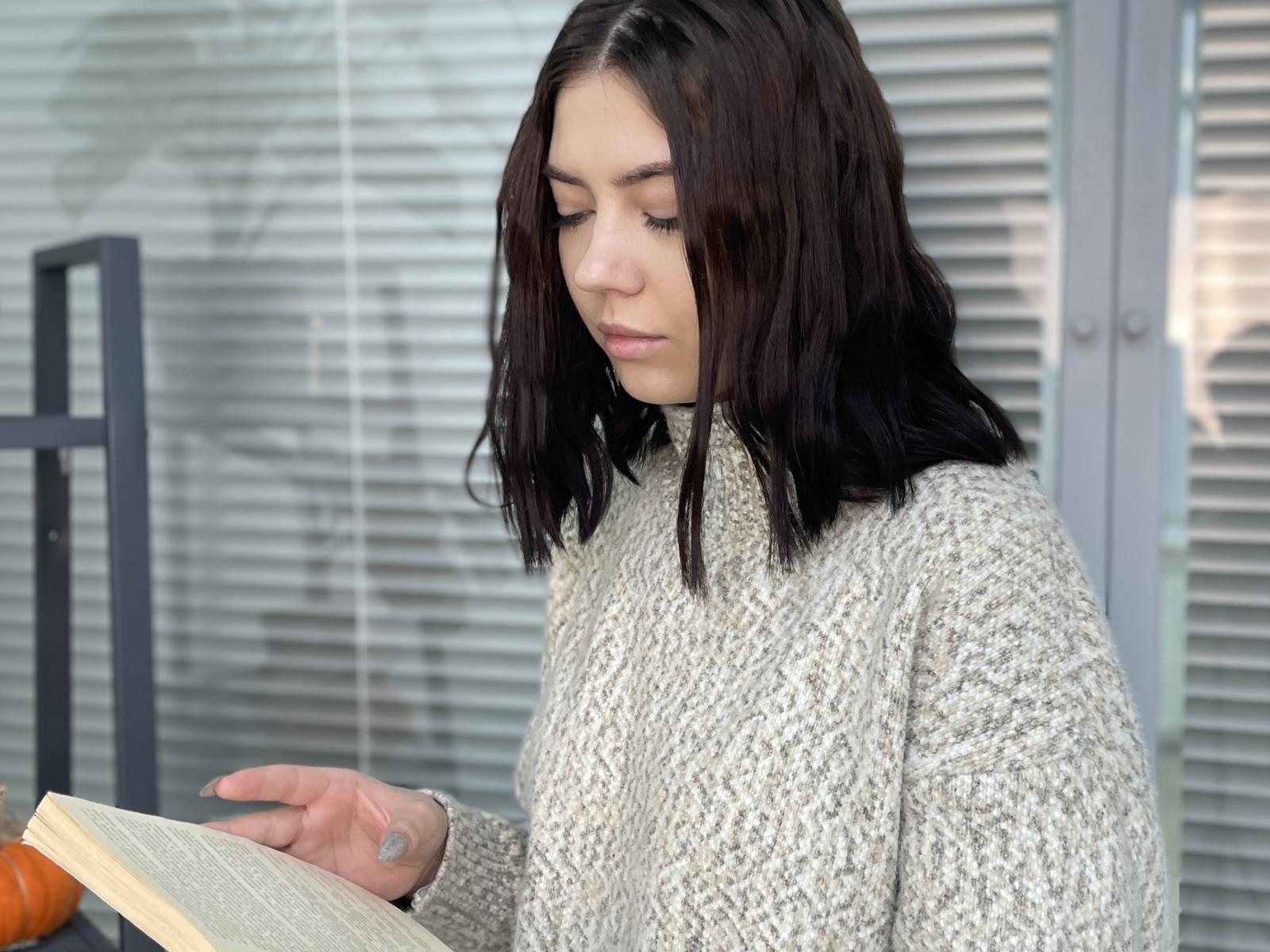 Teresa Lista mit dunkelbraunen Haaren, die einen grauen Strickpullover trägt, liest ein Buch, blickt nach unten und hat einen konzentrierten Gesichtsausdruck, sie hält das Buch mit ihrer rechten Hand..