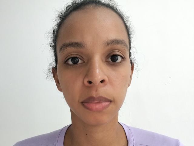 RachelWillians has brown skin, small nose, and curly dark hair in a ponytail. She's wearing a lilac shirt, with neutral expression and pink lips. She's staring straight at camera.