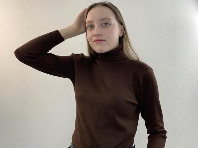 RozanneBarbaria mit heller Haut und blauen Augen, trägt einen braunen Rollkragenpullover, eine Hand im Haar, wirkt leicht gelangweilt, kein Make-up, steht, blondes Haar, weißer Hintergrund..